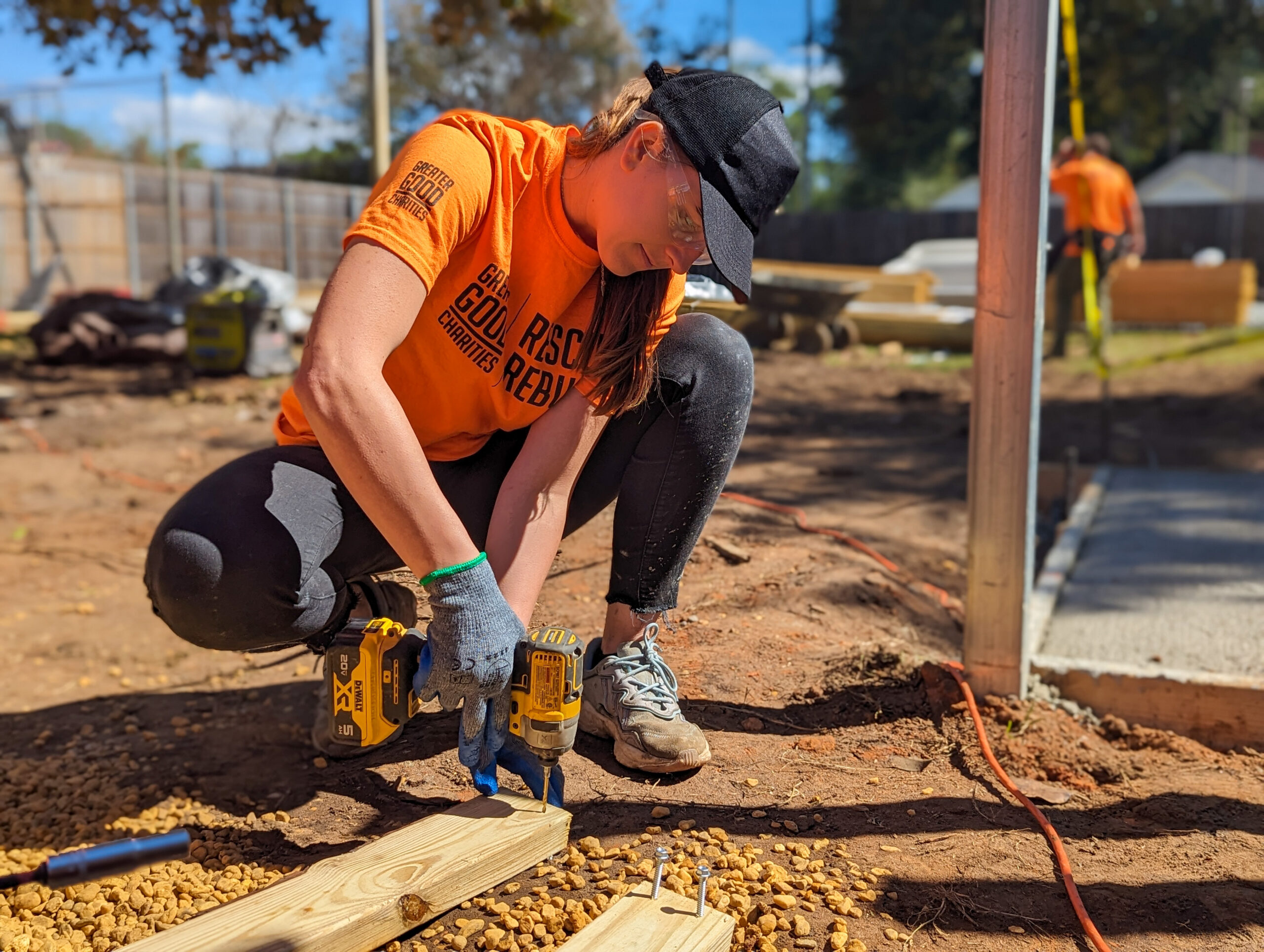 A woman kneels down to drill a hole in a wooden board. Her orange shirt reads "Greater Good Charities Rescue Rebuild".